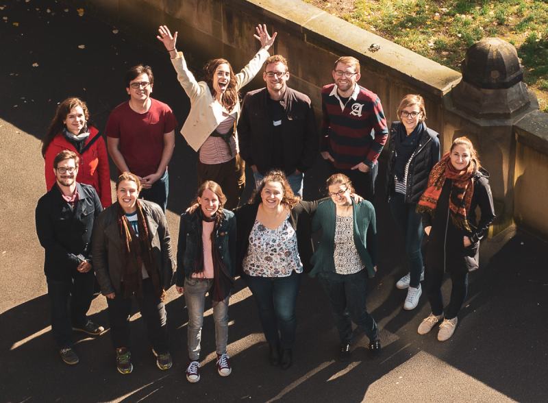 Ein Gruppenportrait aus der Vogelperspektive. Zu sehen sind zwölf Personen im Alter von ca. 20-35 Jahren. Die Chorleiterin hat begeistert die Arme nach oben gestreckt, die anderen haben auch gute Laune.