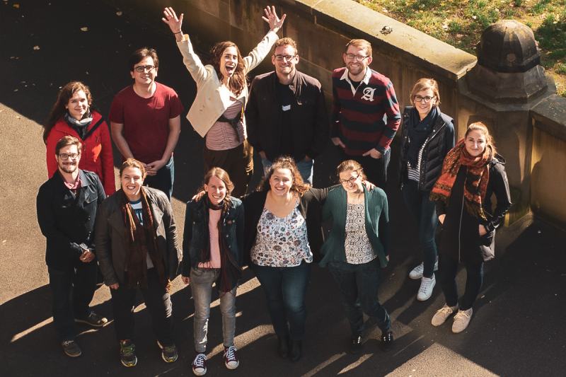 Ein Gruppenportrait aus der Vogelperspektive. Zu sehen sind zwölf Personen im Alter von ca. 20-35 Jahren. Die Chorleiterin hat begeistert die Arme nach oben gestreckt, die anderen haben auch gute Laune.